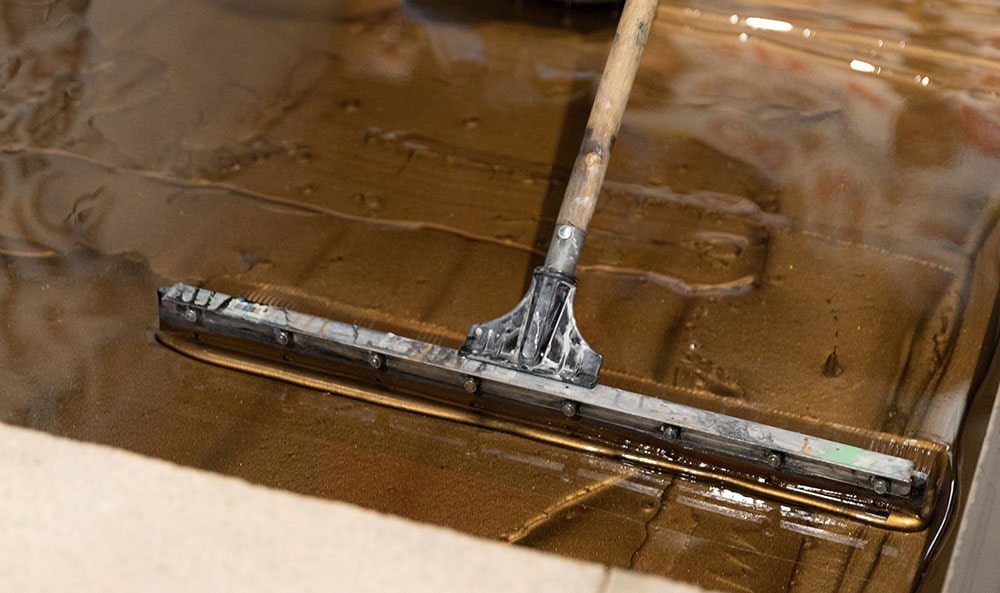 A large squeegee applying a base of epoxy resin flooring over an old concrete foundation.