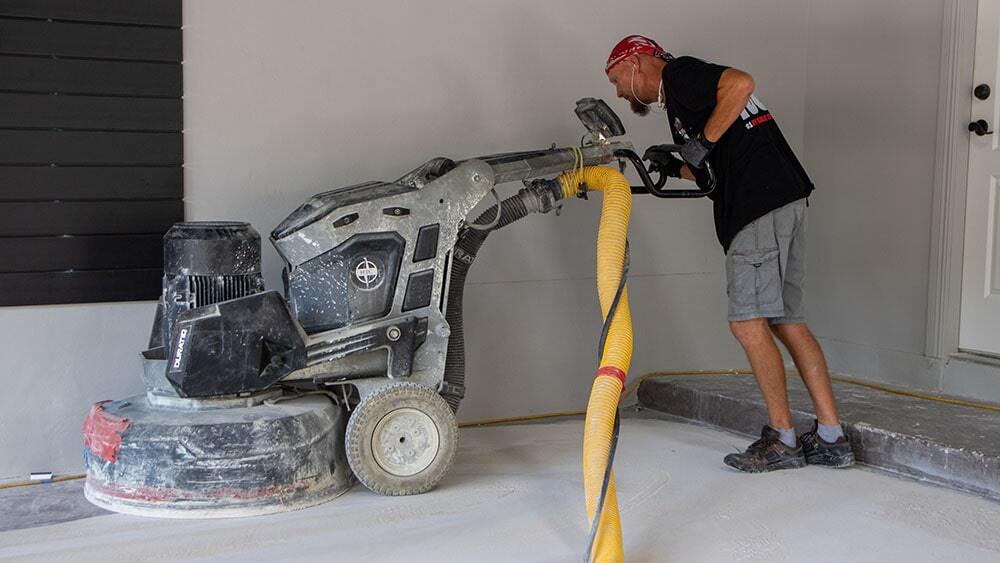 A man is pushing a large concrete grinding machine over a concrete floor in a residential garage.